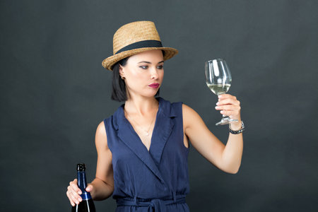 Beautiful young model in a hat tasting wine from the glass while holding a wine bottle with the other hand on a gray background. High quality photo. Studio shot, copy space for youの写真素材