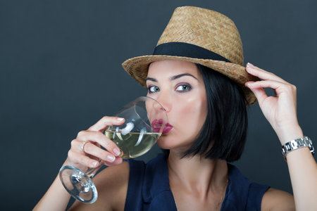 A woman in a sleeveless blue top holds a straw hat and a glass of white wine, with a dark background creating a minimalist, elegant look. Portrait of a beautiful young womenの写真素材