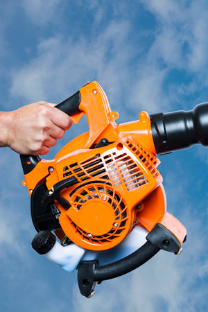 Close up hand holding a Garden blower on blue sky background. Worker with a leaf blower. Landscaper operating gasoline Leaf Blower. Tools. Gardening. Machine. Orange blower machine with black tubeの写真素材