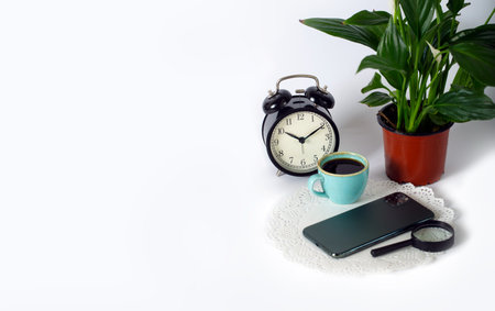Cup of black coffee with alarm clocks, magnifier, phone and Spathiphyllum green houseplant on a...