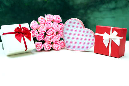 A heart shaped gift box and a red square right box with pink roses placed on a white table.の写真素材