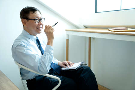 Smart businessman sitting happily thinking on a chair in the office  his hand hold the pen with more copy space.の写真素材