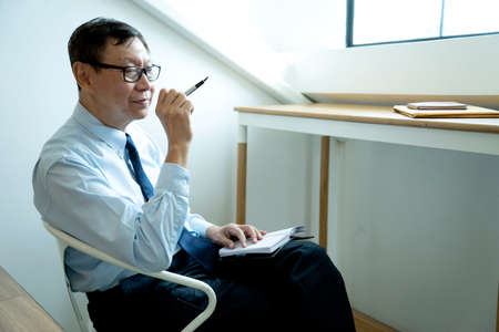 Smart businessman sitting happily thinking on a chair in the office  his hand hold the pen with more copy space.の写真素材