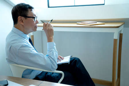 Smart businessman sitting happily thinking on a chair in the office  his hand hold the pen with more copy space.の写真素材
