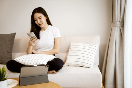 Asian woman sitting on the sofa hold a smart mobile phone and turned to the side with a friendly smile.の写真素材