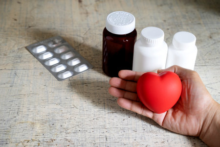 A red heart symbol in his hand, with several pill bottles and a pill box resting on an old white table.の写真素材