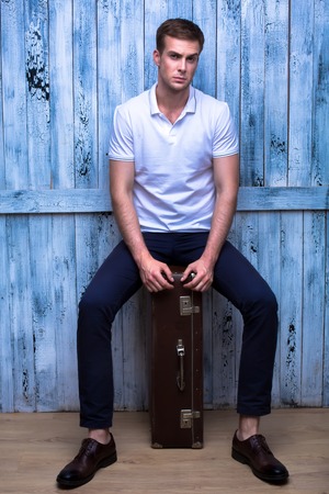 Tall handsome guy in a white polo shirt sitting on an old brown suitcaseの写真素材