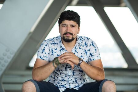 Young bearded man, guy brunette seating inside the bridge construction. Boy, seating in front of the...