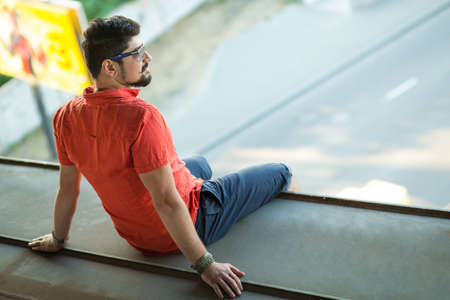 Stylishly dressed guy sits on the bridge dangling his legs and looks away. Bearded young man in glasses and red t-shirtの写真素材