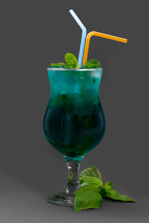 Cocktail with mint and ice in blue glass. Served with tubes and basil on gray backgroundの写真素材