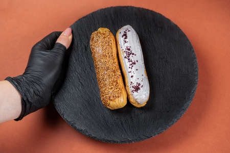 Raspberry and caramel eclair on round black rock stand held by hand in black glove. Eclairs with glaze in cafe. View from above. Cake with choux cream on slate stand. Tasty dessert concept.の写真素材