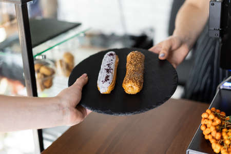 Raspberry and caramel eclair on round black rock stand in hands. Seller gives order to buyer. Sweets to order concept. Two eclairs with white and brown glaze in cafe. Tasty dessert concept.の写真素材