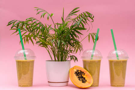 Papaya smoothie with halved papaya and plant pot on pink background. Three papaya smoothies in...