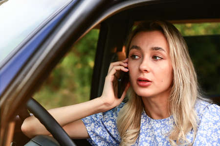Young woman talking on a cell phone while sitting in the car in the drivers seat. Attractive blonde...