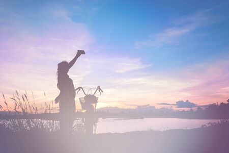 Asian girl taking picture with bike by mobile phone at lake on sunset. Fill color filter.の写真素材