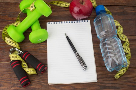Workout and fitness dieting copy space diary. Healthy lifestyle concept. Apple, dumbbell, water, expander hand, stopwatch and measuring tape on rustic wooden table.の写真素材