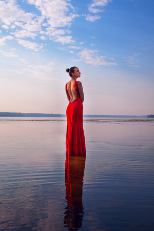 Attractive girl in a red dress standing in water on a background of beautiful sunrise. Beauty, fashion, outdoorの写真素材