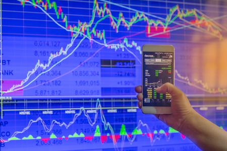 business hand hold and touch screen on smartphone or cellphone over blur stock chart market monitor background,business stock conceptの写真素材