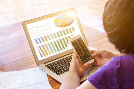 Close-up business woman working in an office interior on a computer holding a smartphone and looking...