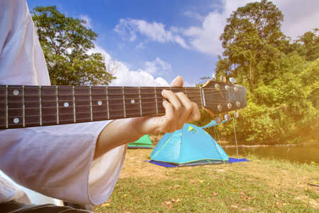 man camping and strum a guitar instrumental music to relax against the background of creek,tent and...