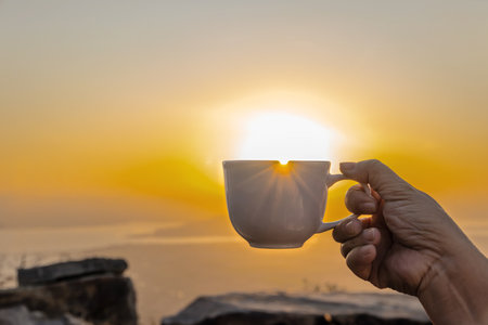Hand holding a white cup of hot espresso coffee mugs and nature view of the sun background in the morning with sunlightの写真素材