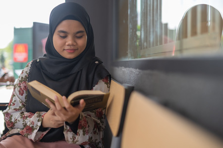 Young cute Muslim Asian woman sitting and reading a book, while chilling and relaxing at restaurant. Student life, study for education concept.の写真素材