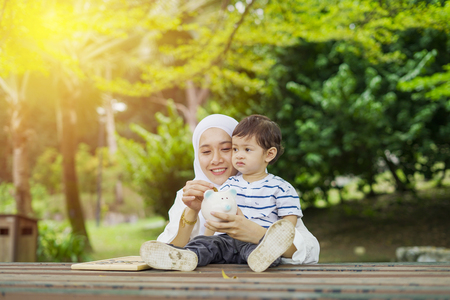Muslim Asian young mother with her baby son, learning and putting coins into piggy bank at park. Education, financial and future planning concept.の写真素材