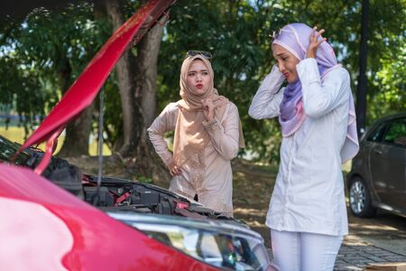 Portrait of two frustrated girls checking front car boot, engine. Car breakdown at side road park, calling for help. Selective focus, copy space.の写真素材