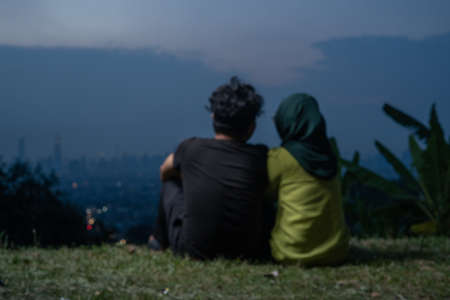 Portrait of Young Asian couple, man and woman, sitting on grass enjoying Kuala Lumpur view during blue hour.の写真素材