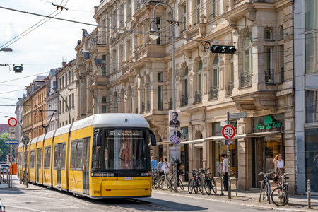 Germany Berlin May 17, 2024. A yellow and black train moves along the street in front of the building. Several people are walking and riding bicycles along the sidewalk.のeditorial素材