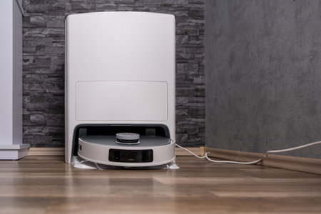 A white robot vacuum cleaner is sitting on a wooden floor. The robot is on a white base and has a black and white design. The robot is not in use and is not movingのeditorial素材