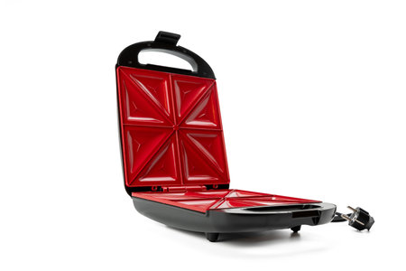 A red and black electric sandwich maker stands on a white background. The electric sandwich maker is...