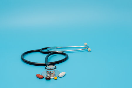 A doctor's stethoscope is on a table with a variety of pills. Scene is serious and focused on the...