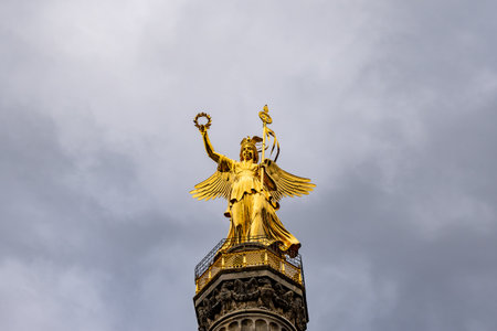 A statue of a woman holding a wreath and a bird. The statue is gold and is on top of a building. The sky is cloudy and the overall mood of the image is serene and peacefulの写真素材