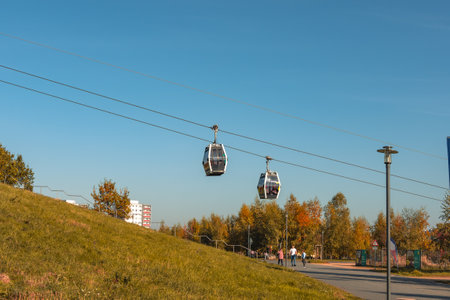 Germany Berlin October 26, 2024.. Two cable cars are suspended in the air above a grassy hill. The sky is clear and blue, and there are several people walking around the area. The scene is peaceful and sereneのeditorial素材