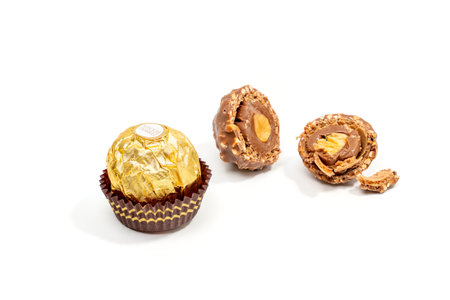 One whole Ferrero Rocher chocolate candy and one cut in half lie on a white background. A chocolate-covered nut with a gold wrapper lies on a white background.のeditorial素材