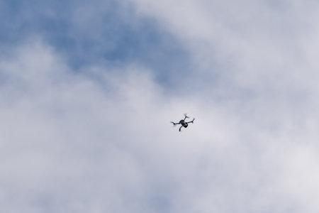 A drone is flying in the sky. The sky is cloudy and the drone is flying high. The drone is black and...
