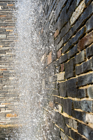 A wall with a stone wall and a water fountain. The water is falling from the top of the fountain
