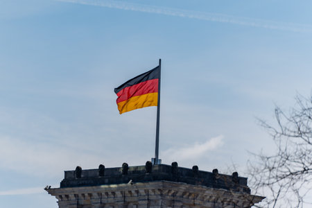 A flag with red, white and black colours flutters on a pole. The German flag is on the roof of the Reichstag building.の写真素材