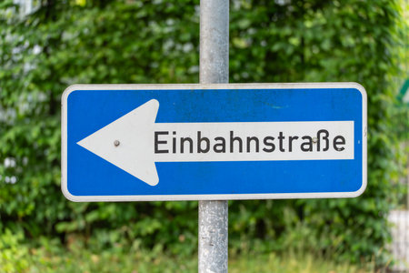 A street sign indicates a one-way street to the left. The sign is set against a backdrop of green foliage.の写真素材