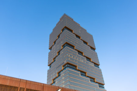 A modern, stacked high-rise tower rises into the blue sky. The facade consists of symmetrical glass...