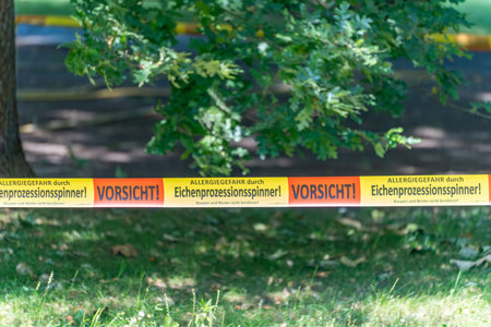 Oak Processionary Moth Warning Tap. A warning tape indicates the danger posed by oak processionary moths. The surrounding area shows oak trees and a sunny park landscape.の写真素材
