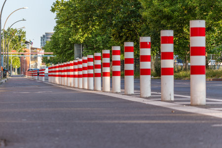 White bollards with red stripes stand in a straight line along a street. This photo shows an urban...