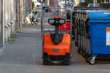Electric pallet jack parked on a sidewalk next to blue and black garbage bins. An everyday urban...