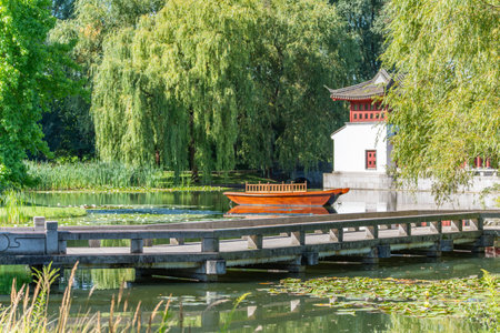 Chinese boathouse with wooden boat. A traditional Chinese building with a red wooden boat is reflected in the calm water. The scene is surrounded by lush greenery and water lilies.の写真素材