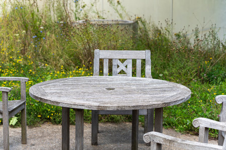 A weathered wooden garden table with matching chairs stands on a patio. Wildflowers and grasses grow...