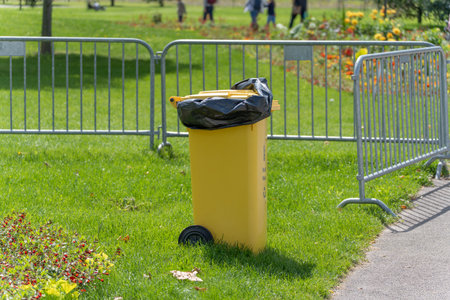 A bright yellow waste bin with a black bag stands on a grassy lawn, fenced by metal barriers. Flowers and people appear in the background.の写真素材