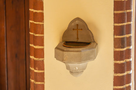 A holy water font with a golden cross is mounted on a church wall. Soft light creates a warm and...