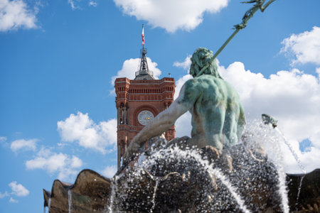 Neptune Fountain Berlin with Red Town Hall. The Neptune Fountain in Berlin sprays water in front of the Red Town Hall. The view is directed towards the striking clock tower.の写真素材
