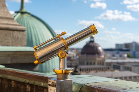 A shiny telescope with golden details stands in front of a green dome. Roofs and the sky are visible in the background.の写真素材
